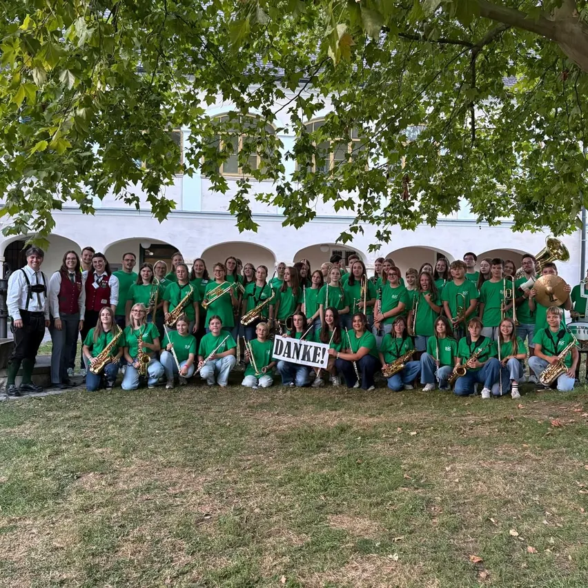 Eine große Gruppe von Menschen in grünen Hemden und Hosen posiert mit Musikinstrumenten vor einem Gebäude für ein Foto. Ein Schild sagt Danke!