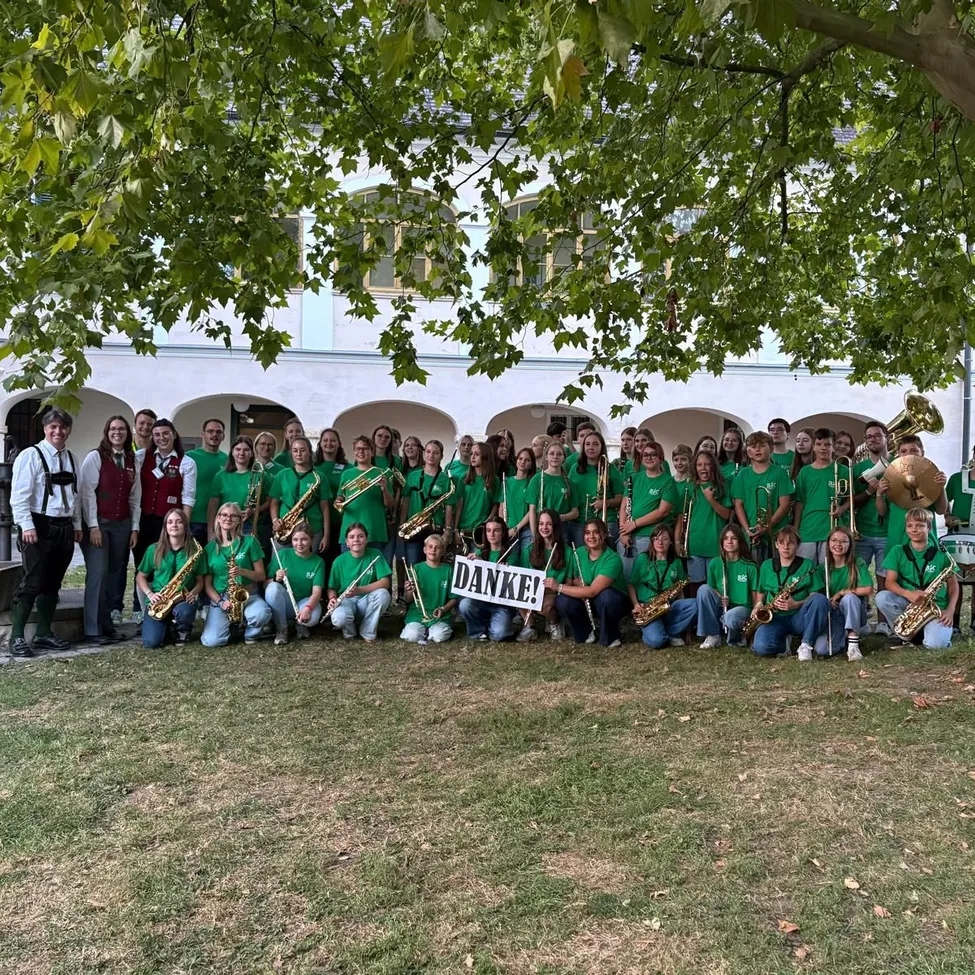 Eine große Gruppe von Menschen in grünen Hemden und Hosen posiert mit Musikinstrumenten vor einem Gebäude für ein Foto. Ein Schild sagt Danke!