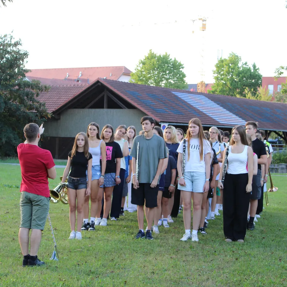 Eine Gruppe junger Menschen, einige mit Musikinstrumenten, steht in einer Reihe auf einem Rasenfeld mit einem Gebäude im Hintergrund.