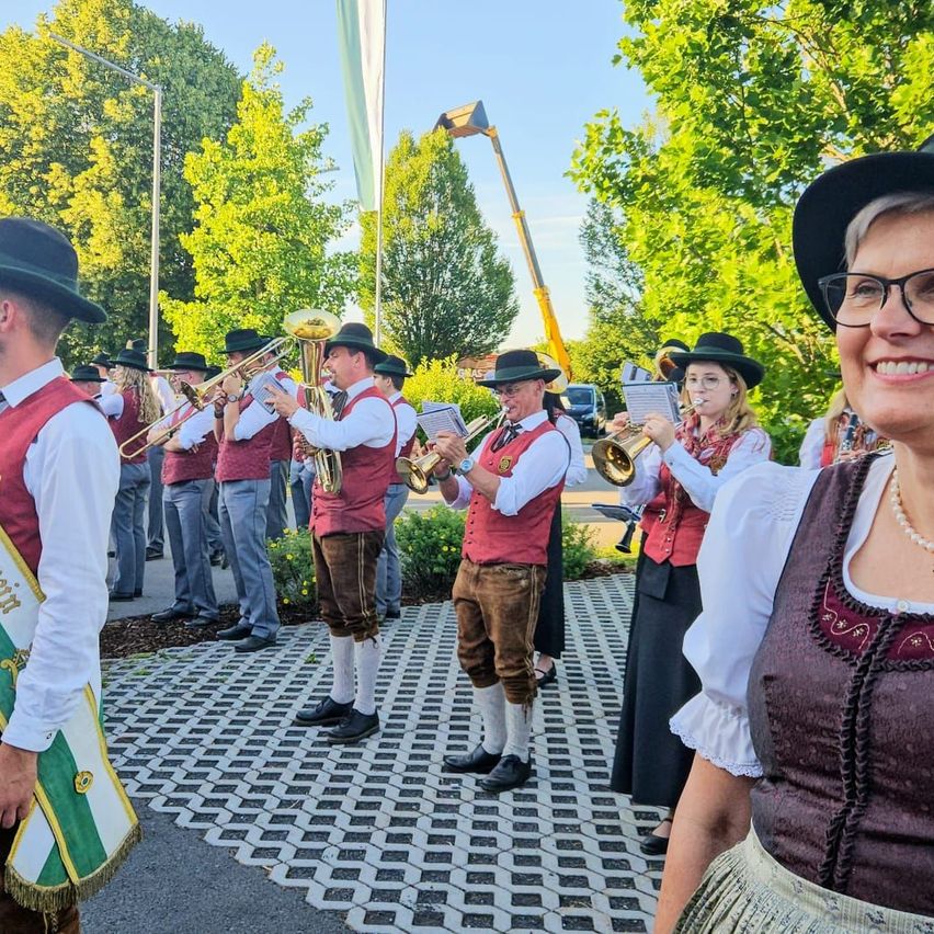 Eine Blaskapelle in traditioneller deutscher Kleidung spielt Instrumente im Freien, umgeben von Grün und einer lächelnden Frau.
