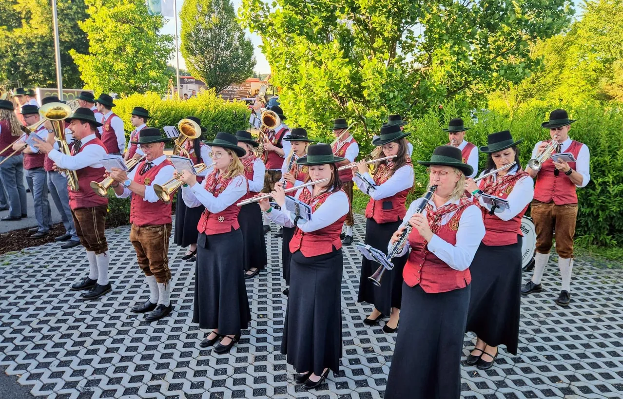 Eine Gruppe von Musikern in traditioneller Kleidung spielt im Freien. Sie spielen verschiedene Instrumente wie Trompeten, Flöten und Posaunen. Sie stehen in einem Park mit Bäumen und Büschen im Hintergrund.