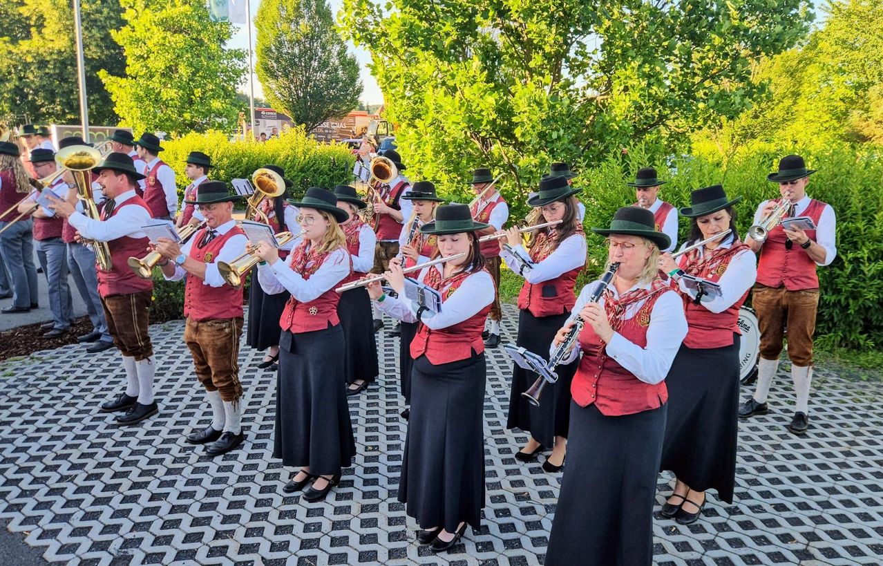 Eine Gruppe von Musikern in traditioneller Kleidung spielt im Freien. Sie spielen verschiedene Instrumente wie Trompeten, Flöten und Posaunen. Sie stehen in einem Park mit Bäumen und Büschen im Hintergrund.