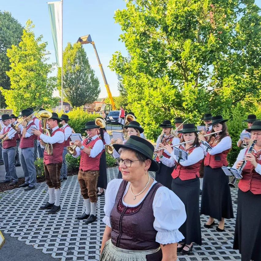 Eine Blaskapelle in traditioneller bayerischer Kleidung steht in einem Außenbereich mit Bäumen und einer Flagge. Die Bandmitglieder spielen verschiedene Instrumente, während eine Frau vor ihnen eine Brille und einen Hut trägt.
