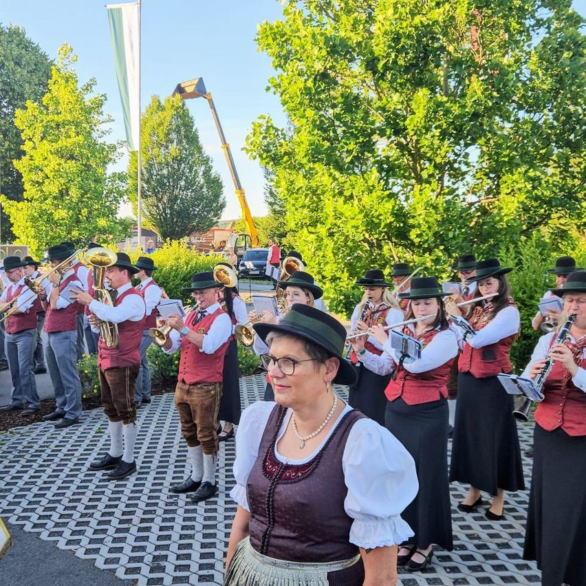 Eine Blaskapelle in traditioneller bayerischer Kleidung steht in einem Außenbereich mit Bäumen und einer Flagge. Die Bandmitglieder spielen verschiedene Instrumente, während eine Frau vor ihnen eine Brille und einen Hut trägt.