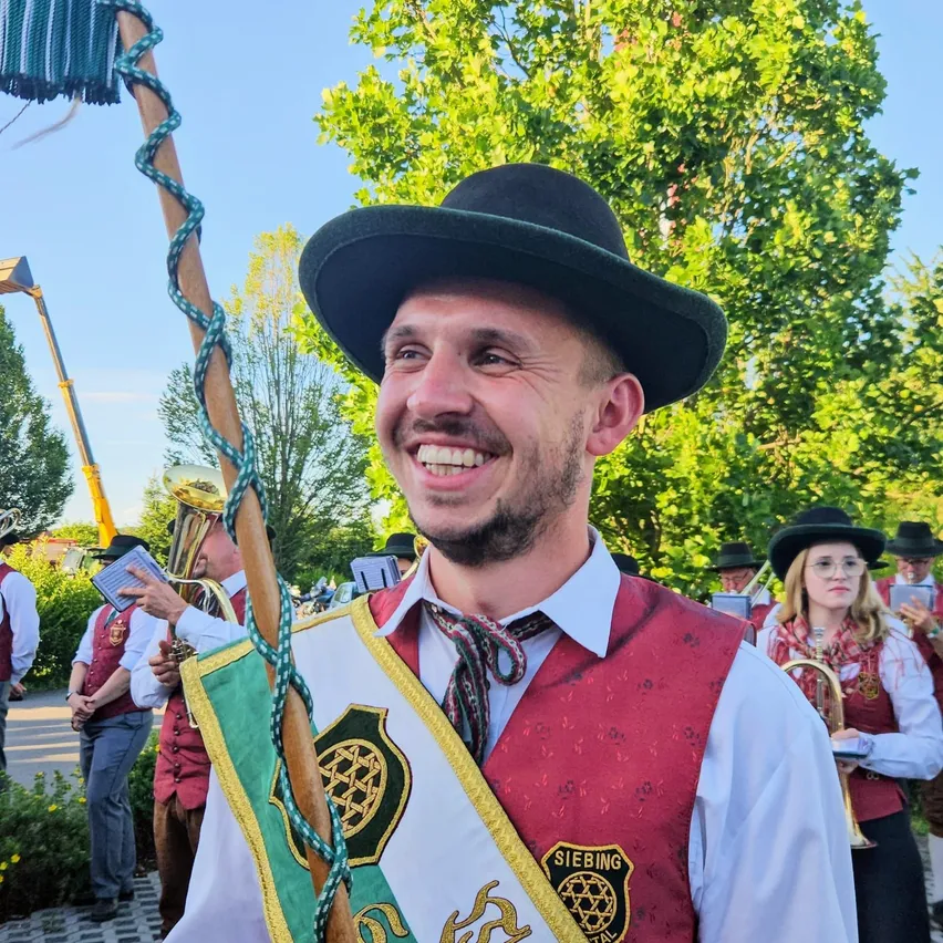 Ein Mann in traditioneller deutscher Kleidung lächelt für die Kamera bei einer Veranstaltung im Freien. Er hält einen Stab mit einer dekorativen Schleife und trägt ein rotes Weste mit einem Logo. Hinter ihm spielen andere in ähnlicher Kleidung Instrumente.