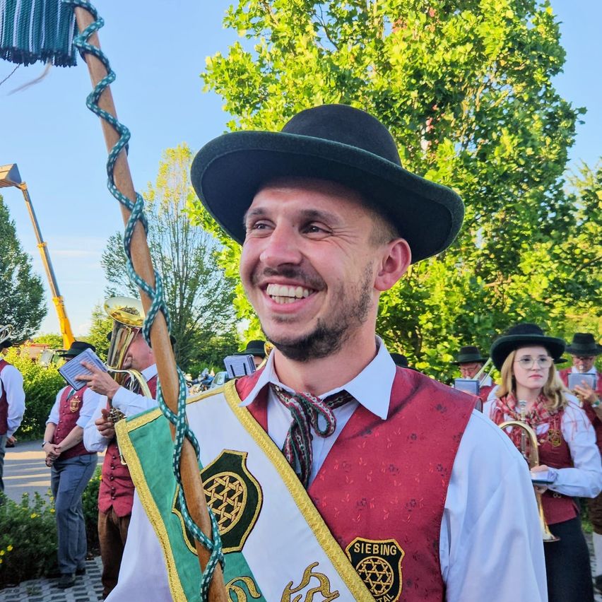 Ein Mann in traditioneller deutscher Kleidung lächelt für die Kamera bei einer Veranstaltung im Freien. Er hält einen Stab mit einer dekorativen Schleife und trägt ein rotes Weste mit einem Logo. Hinter ihm spielen andere in ähnlicher Kleidung Instrumente.