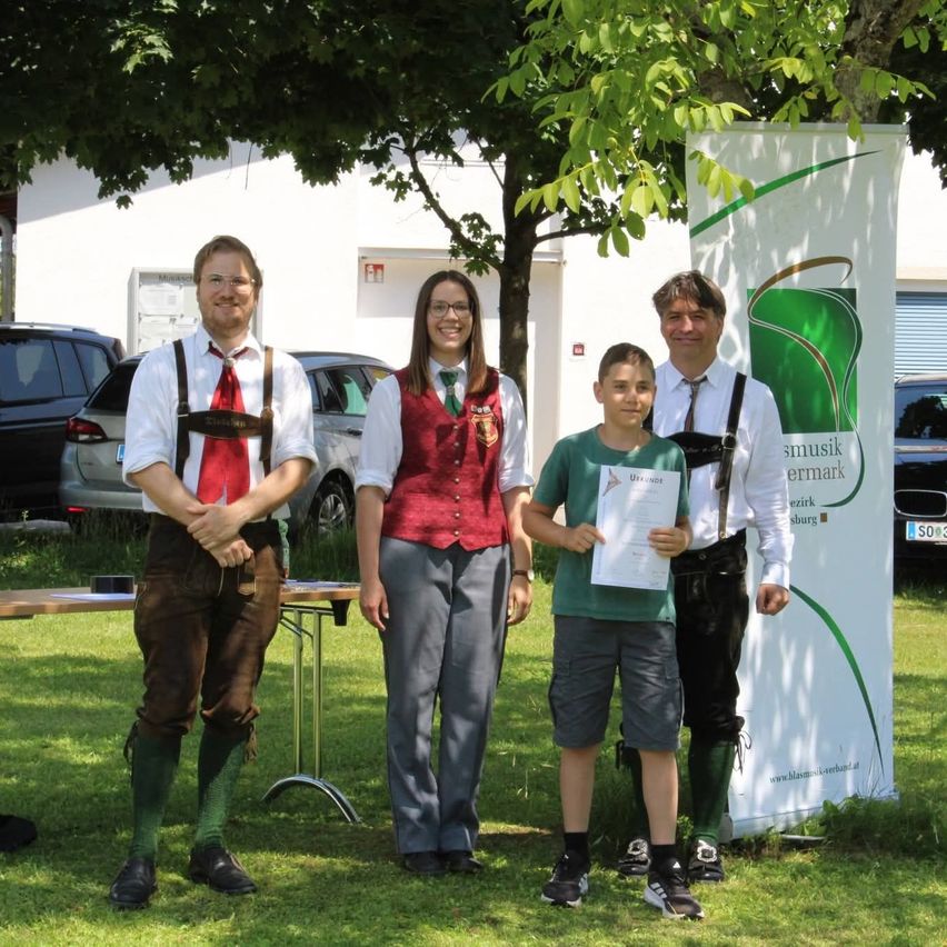 Drei Erwachsene und ein Kind stehen unter einem Baum. Zwei der Erwachsenen tragen traditionelle deutsche Kleidung. Sie alle lächeln. Dahinter steht ein Banner und ein Tisch mit einem geparkten Auto im Hintergrund.