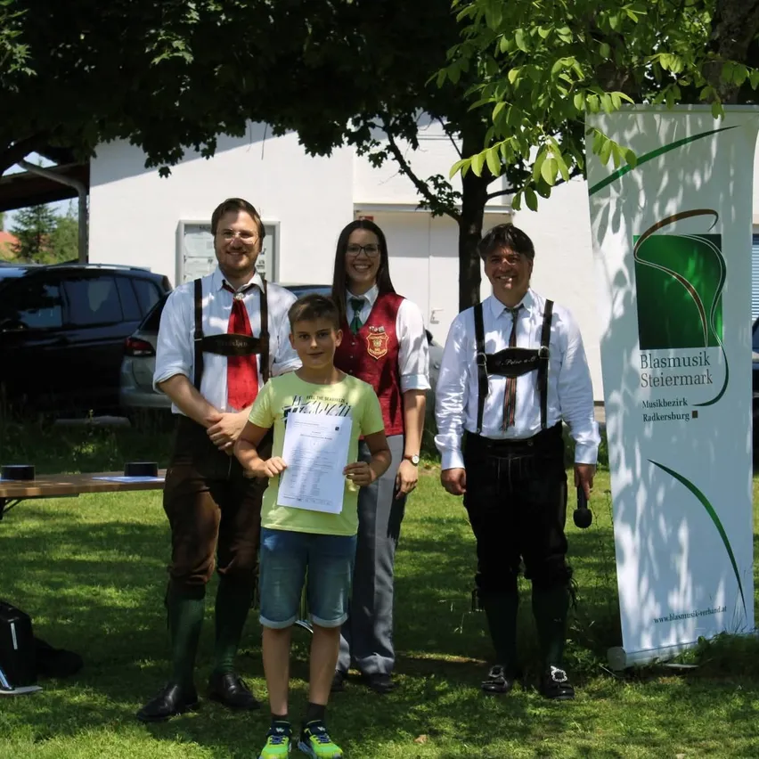 Fünf Personen, drei Männer und zwei Frauen, stehen auf Gras und lächeln. Ein Junge in der Mitte hält ein Papier. Ein Banner steht hinter ihnen.