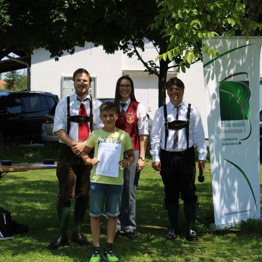 Fünf Personen, drei Männer und zwei Frauen, stehen auf Gras und lächeln. Ein Junge in der Mitte hält ein Papier. Ein Banner steht hinter ihnen.