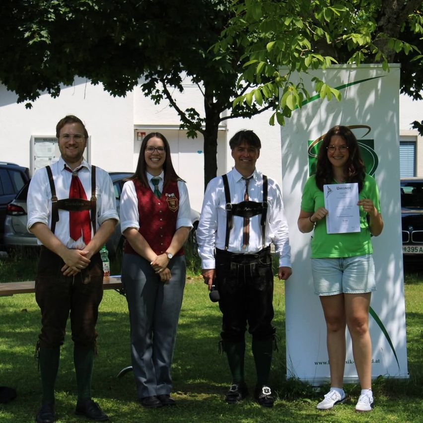 Vier Personen stehen auf einem Rasen, eine Frau hält ein Zertifikat. Hinter ihnen steht eine Frau in einem roten Westchen und lächelt, während zwei Männer in traditioneller deutscher Kleidung neben einem Schild stehen. Eine Bank und geparkte Autos sind im Hintergrund.