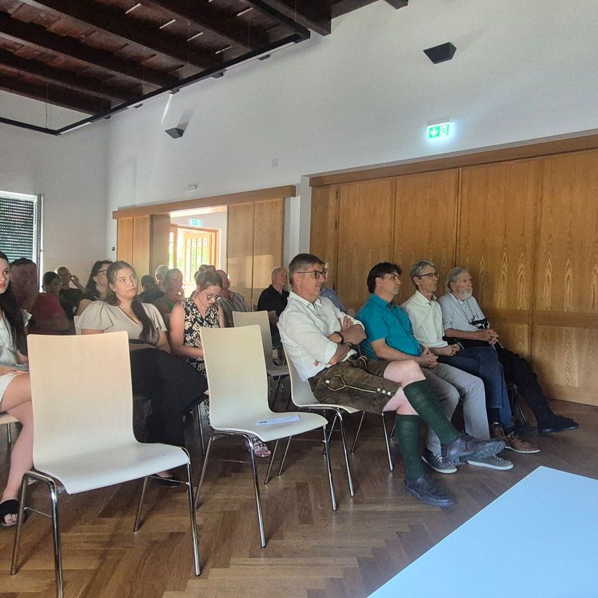 A group of people are sitting in chairs in a room with wooden walls and a wooden floor. Some are wearing glasses and shoes. A blue mat is on the floor.