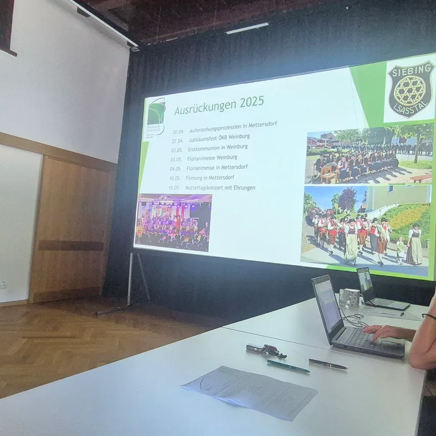 Eine Präsentation auf einem Bildschirm zeigt einen Zeitplan für Veranstaltungen im Jahr 2025 mit Bildern verschiedener Events und Daten. Eine Person sitzt an einem Tisch mit einem Laptop, und ein weiterer Laptop steht auf dem Tisch. Der Raum hat Holzböden und eine weiße Wand.