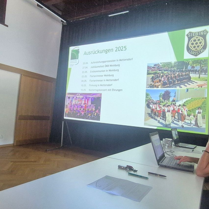 A presentation on a screen shows a schedule for events in 2025, with images of various events and dates. A person is seated at a table with a laptop, and another laptop is on the table. The room has wooden floors and a white wall.