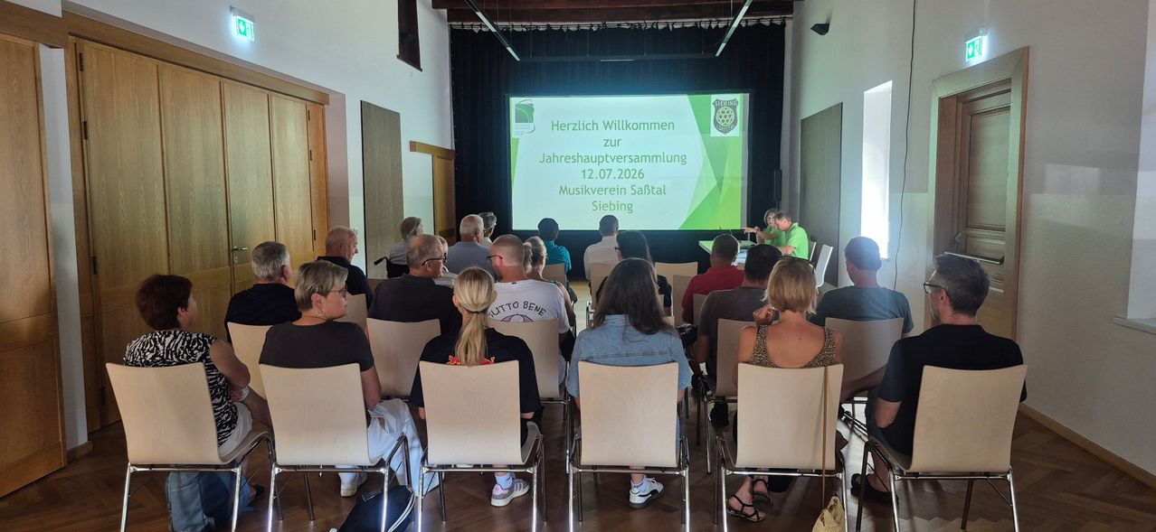 A group of people sit in chairs facing a projector screen displaying a welcome message for the annual meeting of the Musikverein Sabtal Siebing on July 12, 2026.