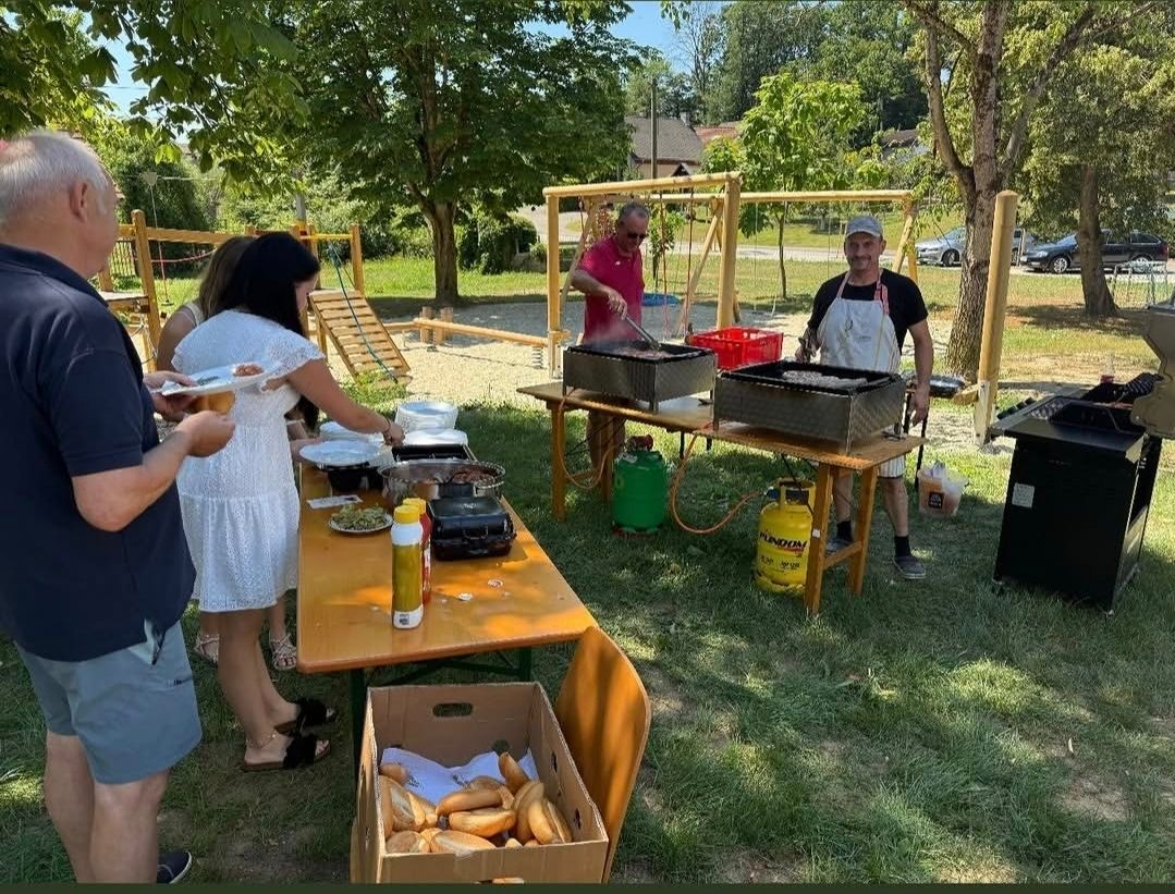 A group of people are preparing food outdoors, with two men grilling on tables and others serving food. There are trees and a playground in the background.