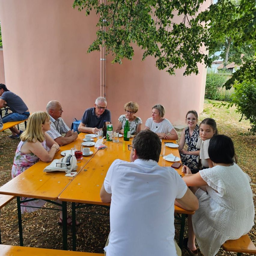 A group of people sitting around a wooden table in an outdoor setting, some with glasses and plates, under a tree with a pink wall in the background.