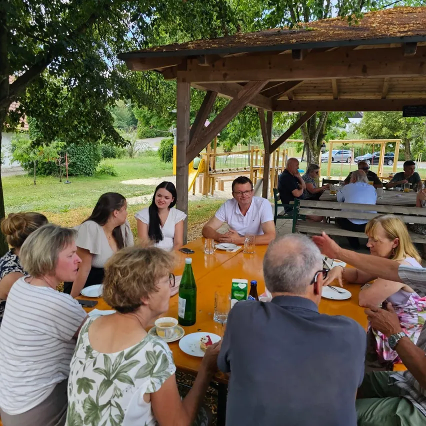 Eine Gruppe von Menschen sitzt um einen Tisch in einem Park, einige unterhalten sich, während andere essen. Ein Mann in einem weißen Hemd schaut eine Frau in einem weißen Oberteil an.