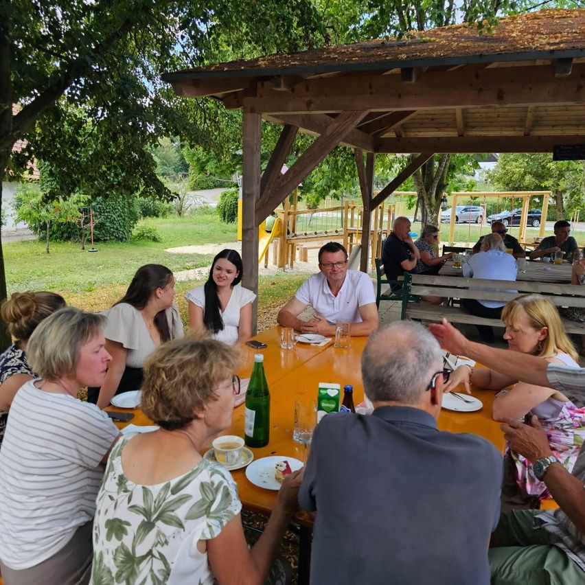 A group of people are sitting around a table in a park, some are talking while others are eating. A man in a white shirt is looking at a woman in a white blouse.