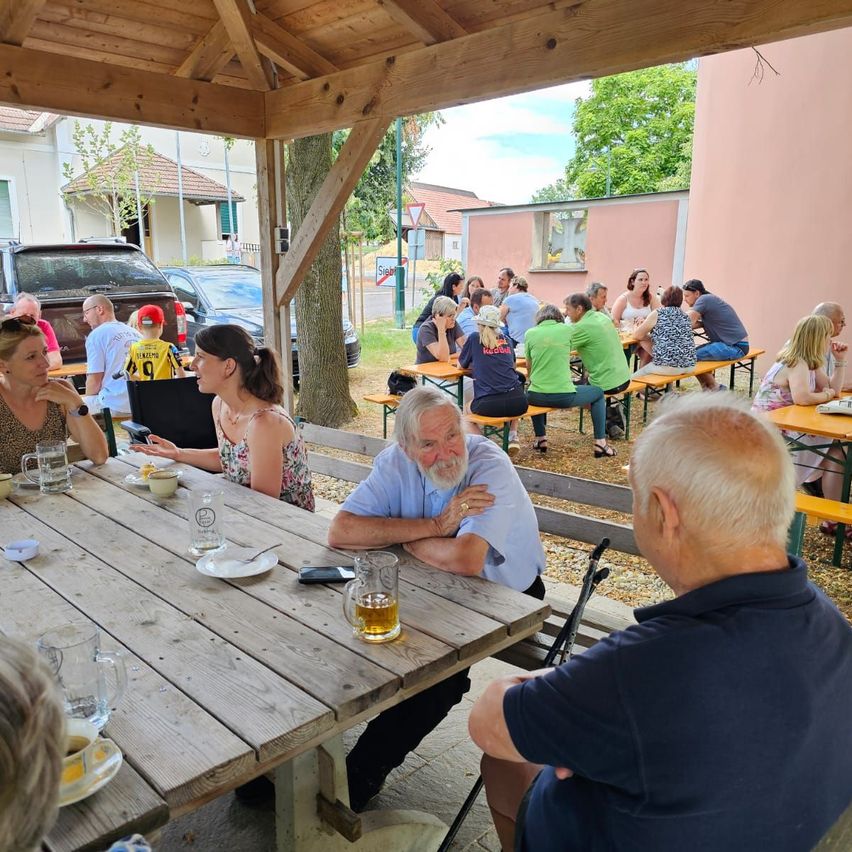 A group of people sit around tables in an outdoor dining area, some wearing cycling jerseys. A man with a beard and mustache holds a glass of beer.