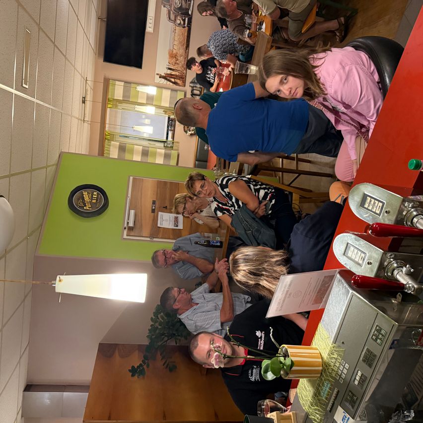 Eine Gruppe von Menschen versammelt sich in einem Restaurant. Einige sitzen, andere stehen. Eine Frau in einem rosa Shirt schaut zur Kamera.