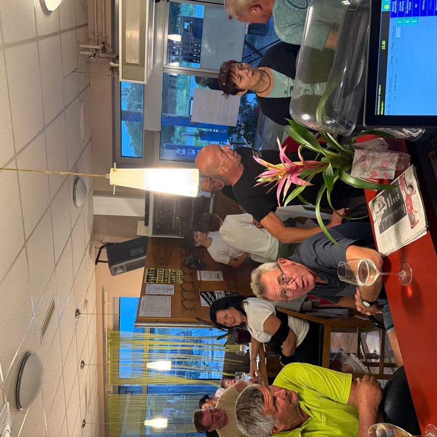Eine Gruppe von Menschen versammelt sich in einem Restaurant, sitzt um einen Tisch mit Weingläsern und einer Blumendekoration. Ein Computerbildschirm steht auf dem Tisch, und die Decke hat Lampen.