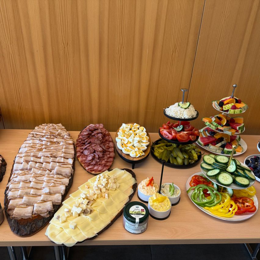 Auf einem Holztisch sind verschiedene Lebensmittel zu sehen, darunter geschnittenes Fleisch, Käse, Eier, Gurken, Früchte und Gemüse, die auf Tellern und Tabletts angeordnet sind.