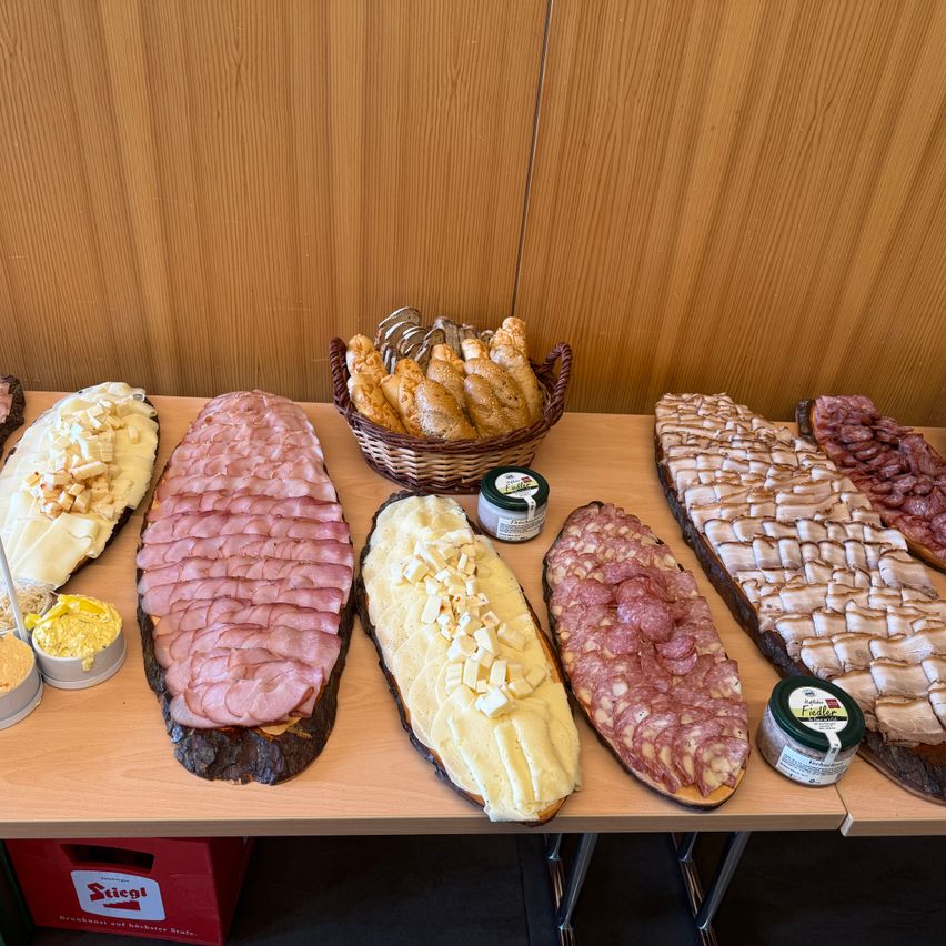 Auf einem Holztisch sind verschiedene Fleisch- und Käsesorten zu sehen, darunter Schinken, Käse und Salami, zusammen mit einem Korb Brot. Die Umgebung deutet auf ein festliches oder gemeinschaftliches Treffen hin.