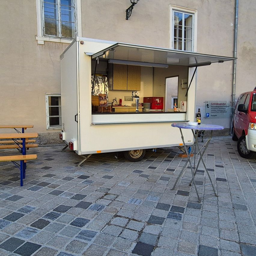 Ein weißer Food Truck ist auf einer Kopfsteinpflasterstraße geparkt, mit offenem Vordach. In der Nähe sind Tische und Bänke. Ein roter Van ist neben dem Gebäude geparkt.