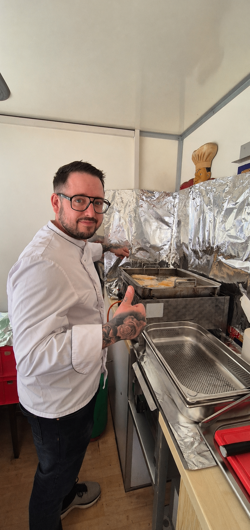 Ein Koch mit Tätowierungen am Arm gibt in einer Küche ein Daumen hoch. Er steht neben einer Speiseschale mit Essen darauf.