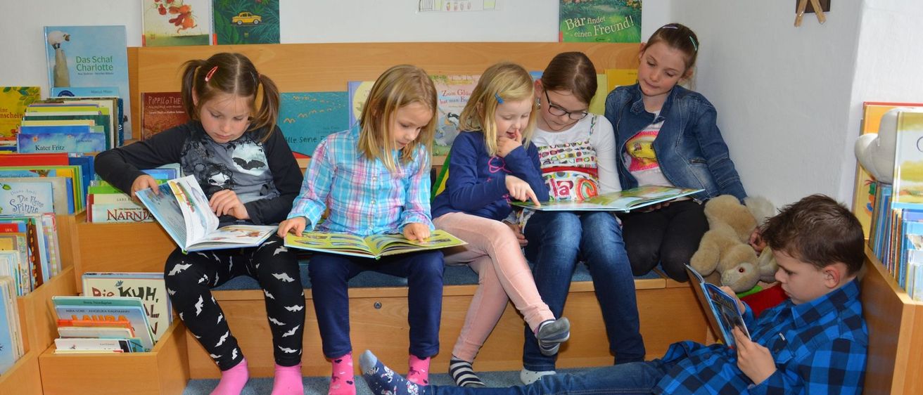 Fünf Mädchen und ein Junge sitzen auf einer Bank und lesen Bücher. Sie sind in einem Raum mit Regalen voller Bücher. Ein Mädchen trägt eine Brille, und ein anderes hat eine Pferdeschwanz. Ein Kuscheltier ist in der Nähe.