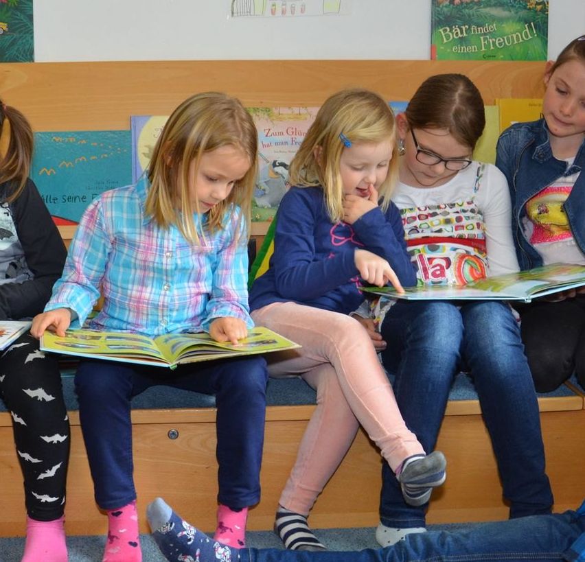 Fünf Mädchen und ein Junge sitzen auf einer Bank und lesen Bücher. Sie sind in einem Raum mit Regalen voller Bücher. Ein Mädchen trägt eine Brille, und ein anderes hat eine Pferdeschwanz. Ein Kuscheltier ist in der Nähe.