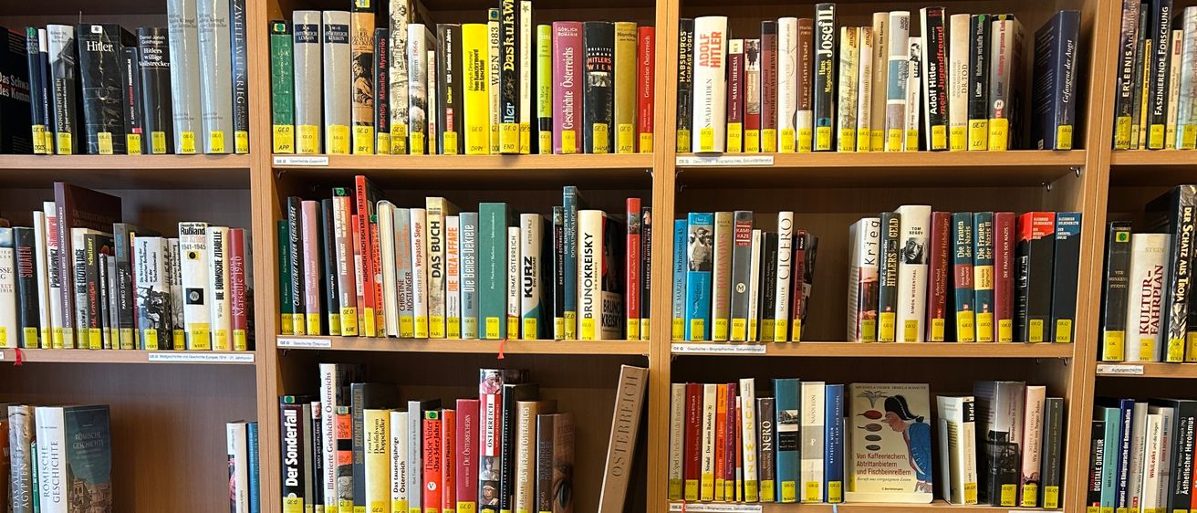 Ein hölzerne Bücherregal voller Bücher, kategorisiert in Bereiche wie 'Gesellschaft, Politik, Medien, Recht, Wirtschaft'. Die Bücher sind ordentlich mit gelben Etiketten angeordnet.