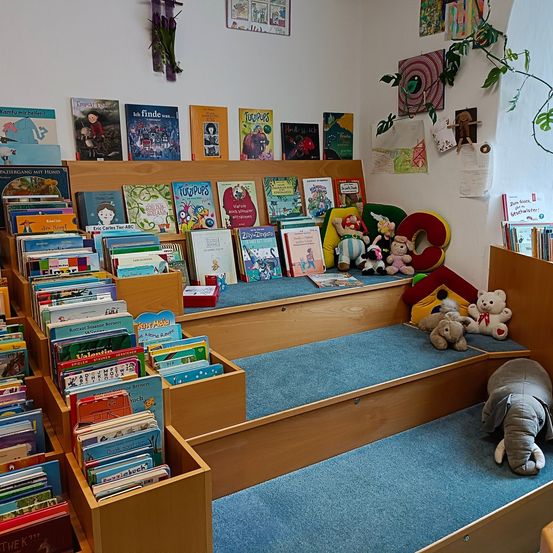 Ein Kinderzimmer gefüllt mit Büchern auf Holzregalen und weichen Teppichen. Gefüllte Tiere und Spielzeug sind verstreut, und bunte Buchstaben des Alphabets sind an der Wand.