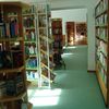 Eine Innenbibliothek mit Reihen von Bücherregalen, gefüllt mit verschiedenen Büchern. Sonnenlicht strömt durch eine Tür und beleuchtet den Gang. Einige Personen sind in der Ferne zu sehen.