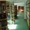 Eine Innenbibliothek mit Reihen von Bücherregalen, gefüllt mit verschiedenen Büchern. Sonnenlicht strömt durch eine Tür und beleuchtet den Gang. Einige Personen sind in der Ferne zu sehen.