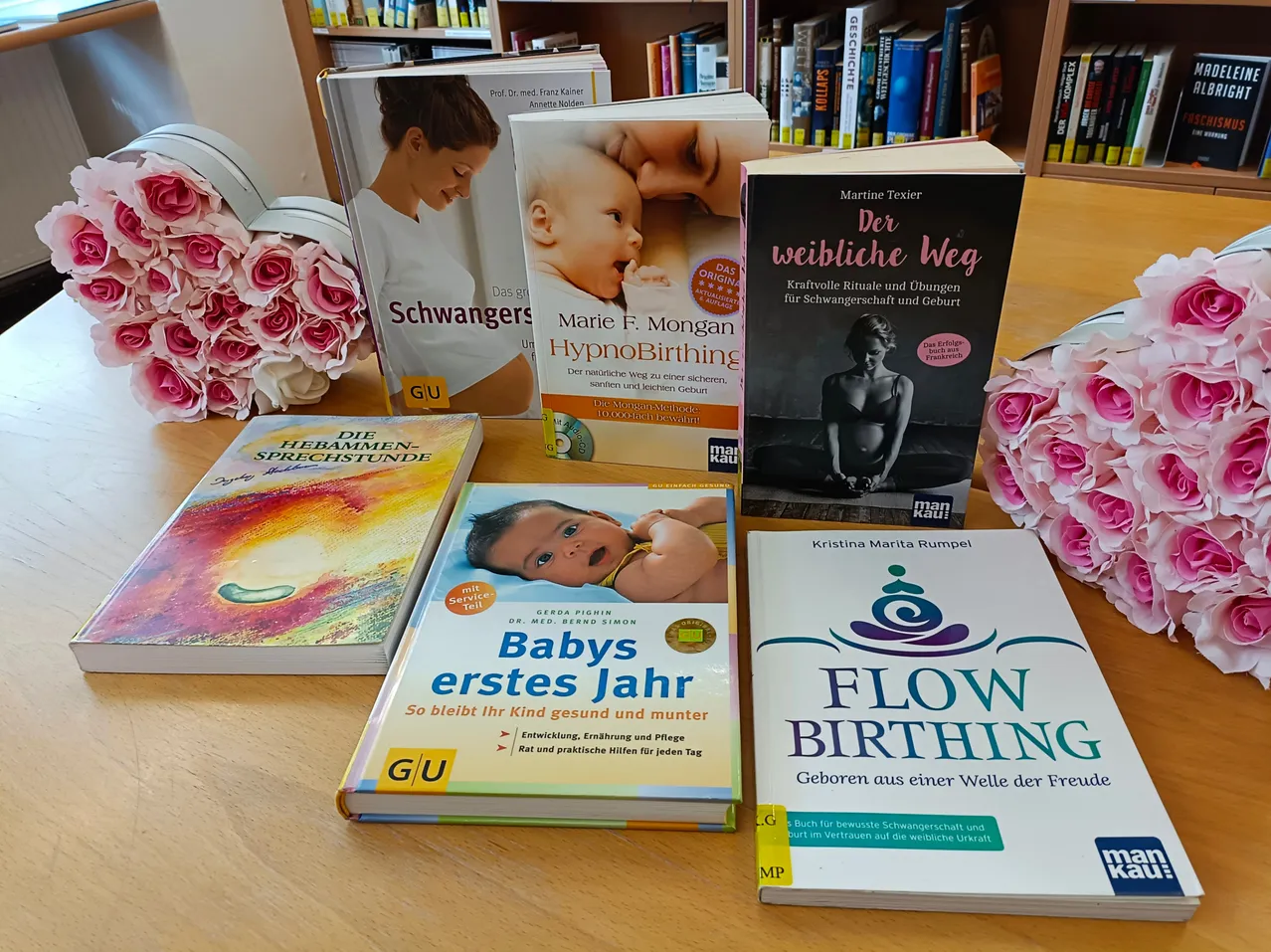 Eine Sammlung von Büchern über Schwangerschaft und Geburt, einschließlich Titel wie 'Schwangerschaft', 'Marie F. Mongan HypnoBirthing' und 'Der weibliche Weg', auf einem Holztisch angeordnet mit rosa Rosen im Hintergrund.