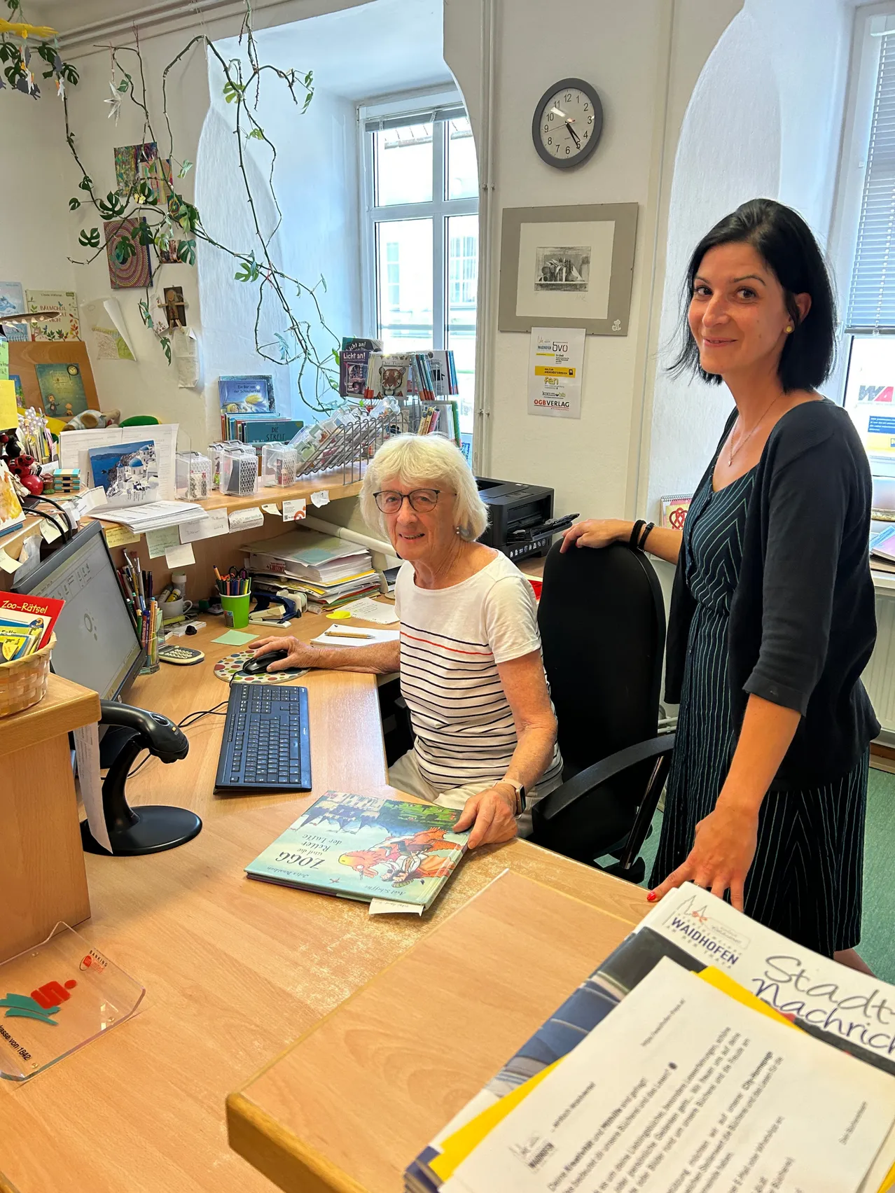 Zwei Frauen sind in einem Büro. Eine Frau sitzt an einem Schreibtisch mit einem Computer und einem Buch, während die andere Frau hinter ihr steht und lächelt.