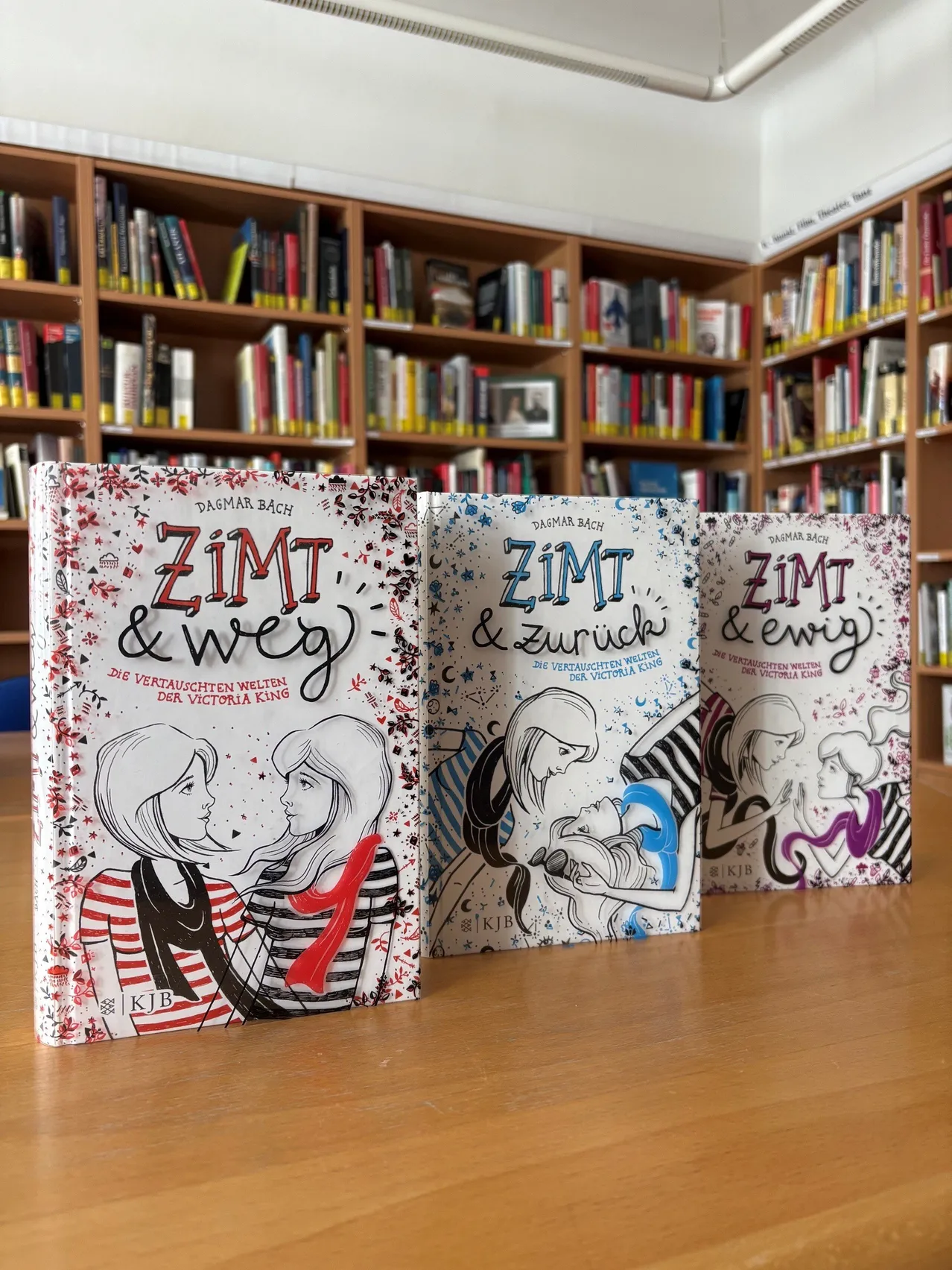 Drei Bücher mit den Titeln 'Zimt & weg', 'Zimt & zurück' und 'Zimt & ewig' sind auf einem Holztisch vor einem Bücherregal mit Büchern ausgestellt.