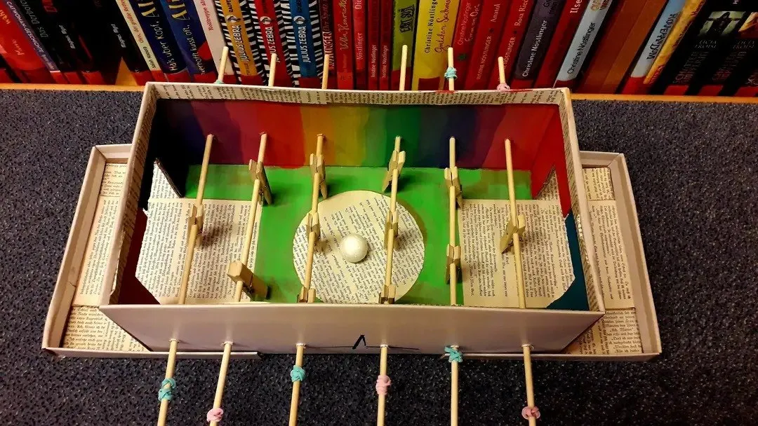 Eine selbstgemachte Tischfußball-Spielfläche aus Pappe und Stöcken, mit einem weißen Ball in der Mitte, auf einer schwarzen Oberfläche mit Büchern im Hintergrund.