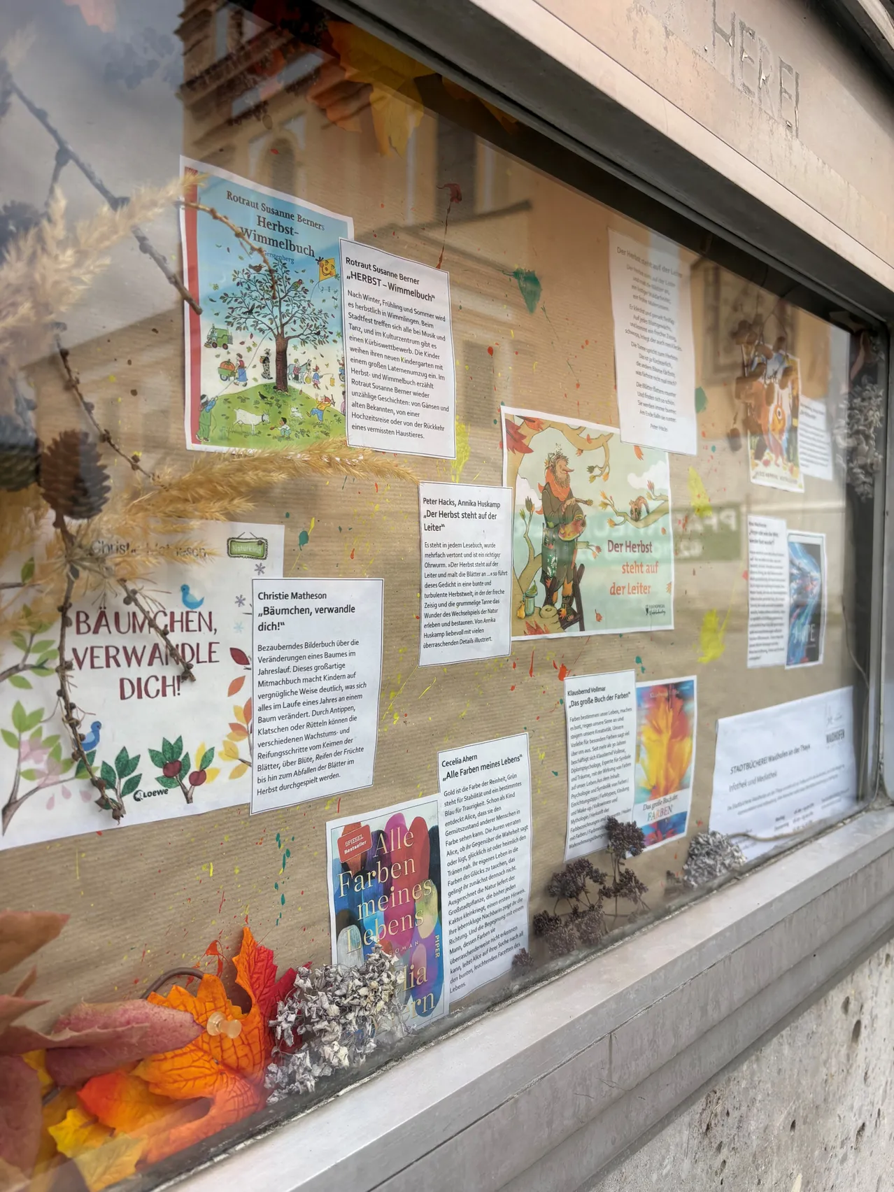 Ein Bulletinboard mit verschiedenen farbigen Plakaten und Papieren, darunter ein Poster mit der Aufschrift 'BAUMCHEN, VERWANDLE DICH!' und ein anderes mit der Aufschrift 'Alle Farben meines Lebens'.
