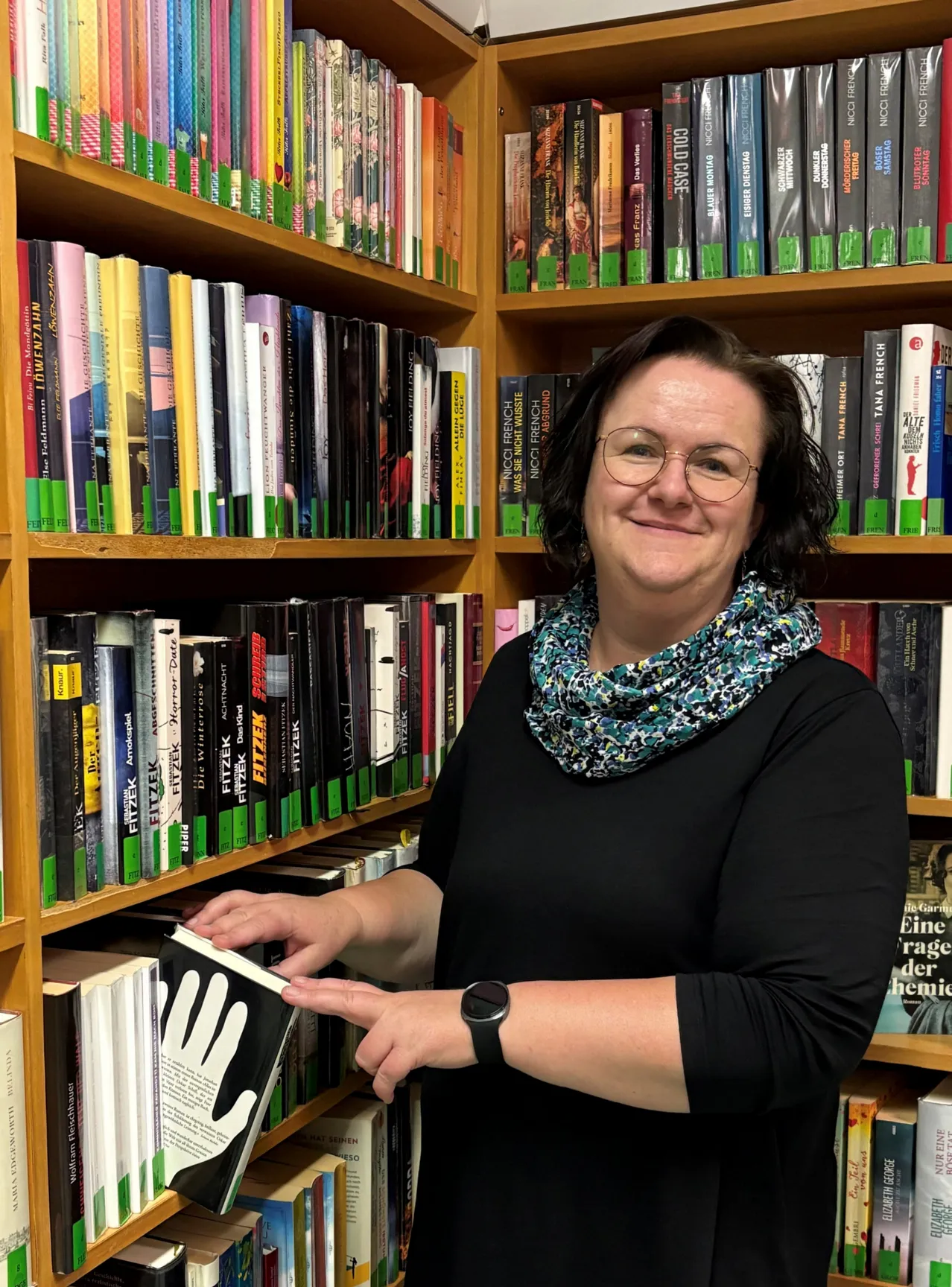 Eine Frau mit Brille steht in einer Bibliothek und hält ein Buch mit einem Handabdruck auf dem Cover.