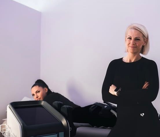 Zwei Frauen in einem Raum. Eine Frau liegt auf einem Bett mit einem Gerät neben ihr. Die andere Frau steht mit verschränkten Armen.