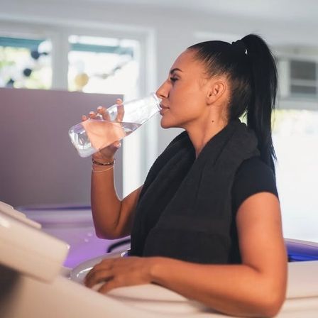Eine Frau trinkt Wasser aus einer Flasche, während sie auf einem Stuhl sitzt, eine schwarze Bluse und Weste trägt, mit einem unscharfen Hintergrund, der ein Fenster und lila Lichter zeigt.