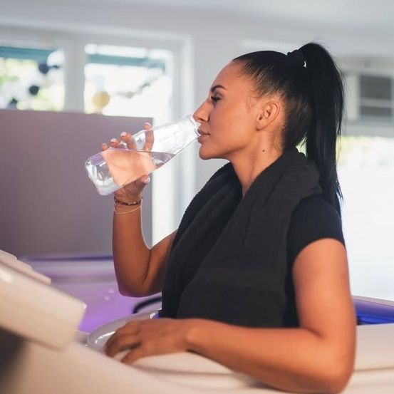 Eine Frau trinkt Wasser aus einer Flasche, während sie auf einem Stuhl sitzt, eine schwarze Bluse und Weste trägt, mit einem unscharfen Hintergrund, der ein Fenster und lila Lichter zeigt.