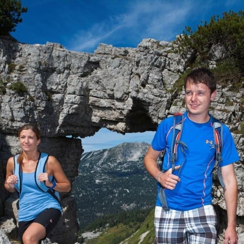 Ein Mann und eine Frau wandern in einem Berggebiet und stehen auf einer Felsformation mit einem natürlichen Bogen. Der Mann trägt ein blaues T-Shirt und karierte Shorts, während die Frau ein blaues Tanktop und schwarze Shorts trägt. Sie haben beide Rucksäcke.