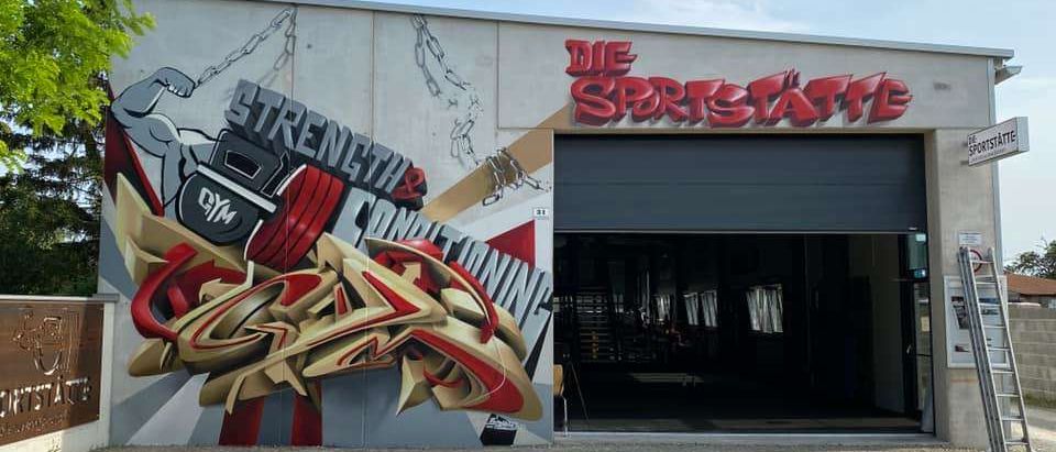 Ein Fitnessstudiogebäude mit einer großen Graffiti-Wandmalerei, die eine muskulöse Figur beim Gewichtheben und den Text 'STÄRKE & KONDITIONIERUNG' zeigt. Über der Wandmalerei ist der Name des Fitnessstudios 'DIE SPORTSTÄTTE' zu sehen. Das Gebäude hat eine Garagentür und einen klaren Blick ins Innere.