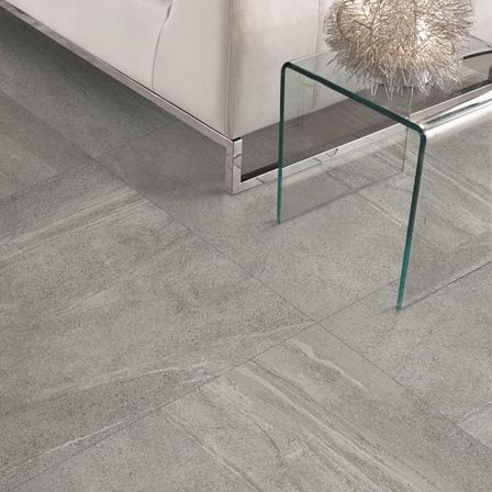 Ein modernes Wohnzimmer mit einer weißen Couch, grau gefliestem Boden und einem Glastisch mit einer dekorativen Lampe.