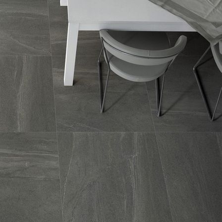 Ein weißer Tisch und zwei graue Stühle stehen auf einem grau gefliesten Boden. Auf dem Tisch liegt ein grauer Stoff mit einer Kerze und Steinen darauf.