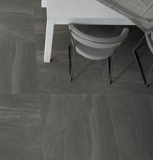 Ein weißer Tisch und zwei graue Stühle stehen auf einem grau gefliesten Boden. Auf dem Tisch liegt ein grauer Stoff mit einer Kerze und Steinen darauf.