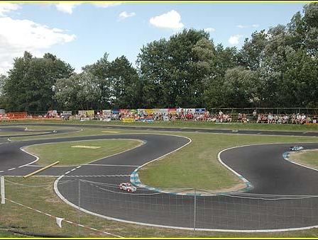 Eine Rennstrecke mit zwei Autos darauf. Ein Publikum beobachtet von den Seitenlinien. Bäume sind im Hintergrund.